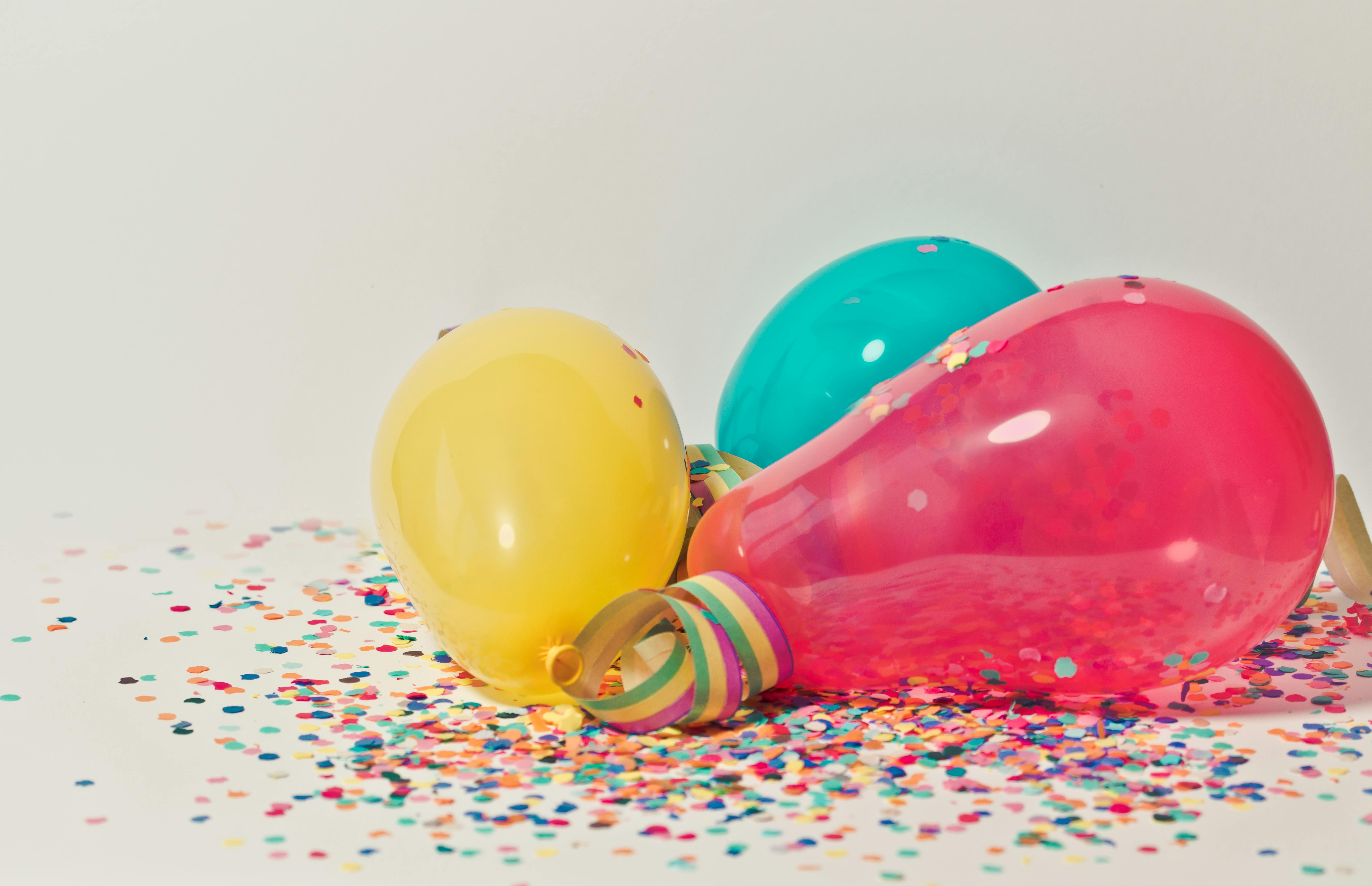 Yellow, pink, and teal balloons on a white bakground surrounded by confetti